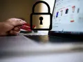 Dłoń trzyma kartę debetową przy laptopie z otwartą stroną sklepu internetowego. Symbol kłódki nad kartą sugeruje, czy kartą debetową można płacić w internecie bezpiecznie.