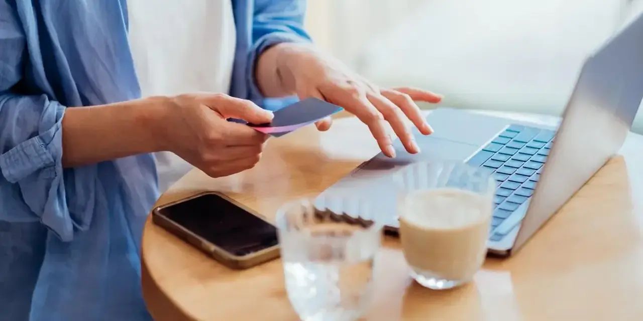 Kobieta dokonuje płatności kartą w internecie, wpisując dane na laptopie. Obok leży telefon i napoje.