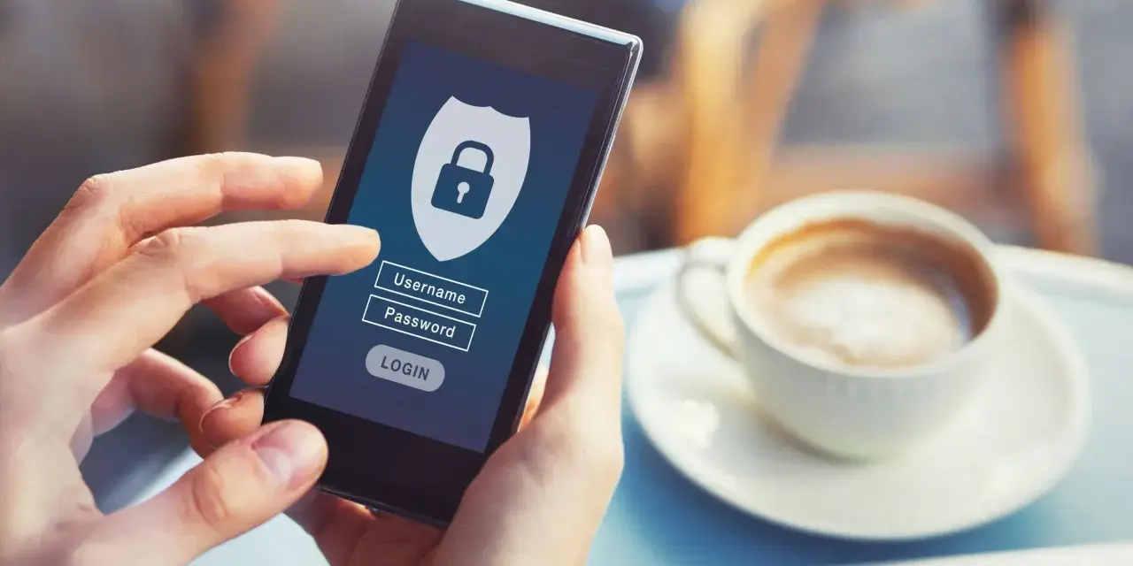 Dłoń trzyma smartfon z ekranem logowania do banku. Widoczny symbol tarczy z kłódką, pola na login i hasło.