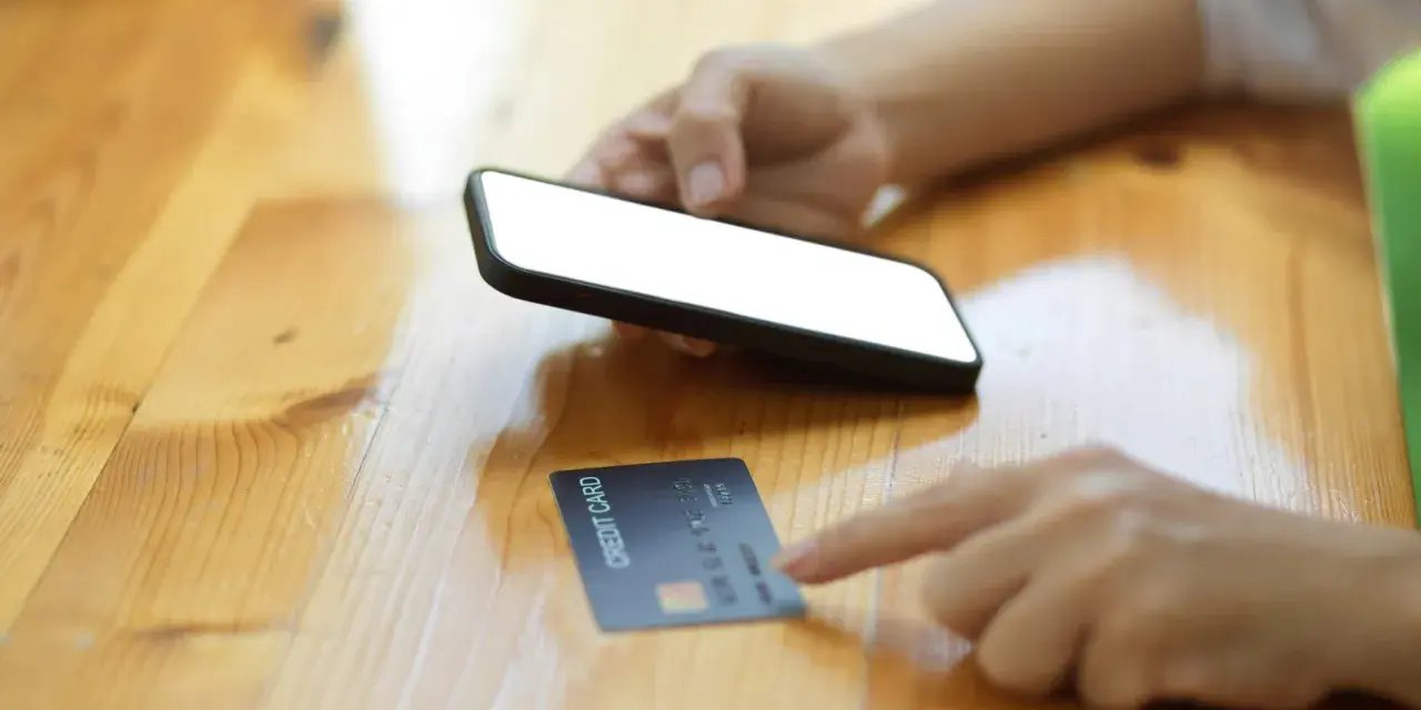 Dłoń trzyma smartfon, obok leży karta kredytowa. Szybkie zakupy online, jak w millennium impresja sklepy.