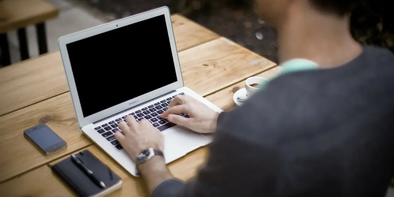 Mężczyzna pracuje na laptopie przy stoliku na zewnątrz. Obok leży telefon, notatnik i kawa. Może właśnie zarabia na **moneyo.pl**.