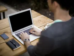 Mężczyzna pracuje na laptopie przy stoliku na zewnątrz. Obok leży telefon, notatnik i kawa. Może właśnie zarabia na **moneyo.pl**.