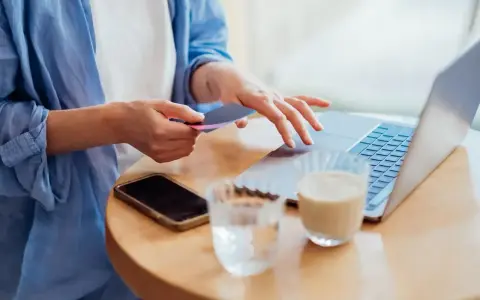 Kobieta dokonuje płatności kartą w internecie, wpisując dane na laptopie. Obok leży telefon i napoje.