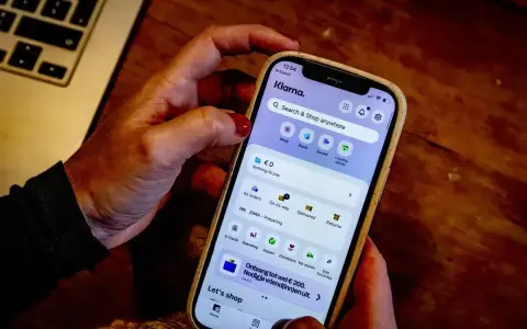 Dłoń trzyma smartfon z aplikacją Klarna. Czy Klarna jest bezpieczna? Aplikacja oferuje zakupy, płatności i zarządzanie zamówieniami.