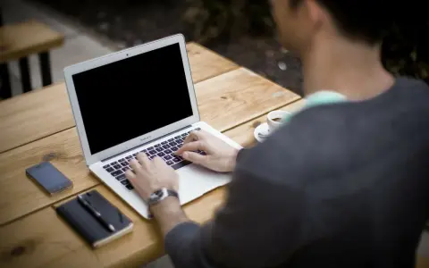 Mężczyzna pracuje na laptopie przy stoliku na zewnątrz. Obok leży telefon, notatnik i kawa. Może właśnie zarabia na **moneyo.pl**.
