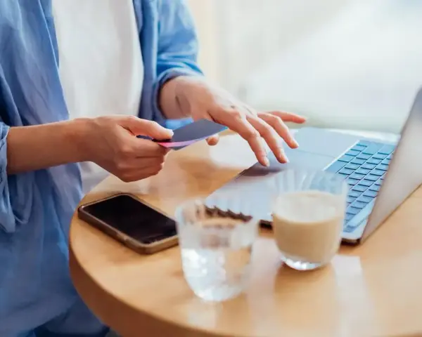 Kobieta dokonuje płatności kartą w internecie, wpisując dane na laptopie. Obok leży telefon i napoje.