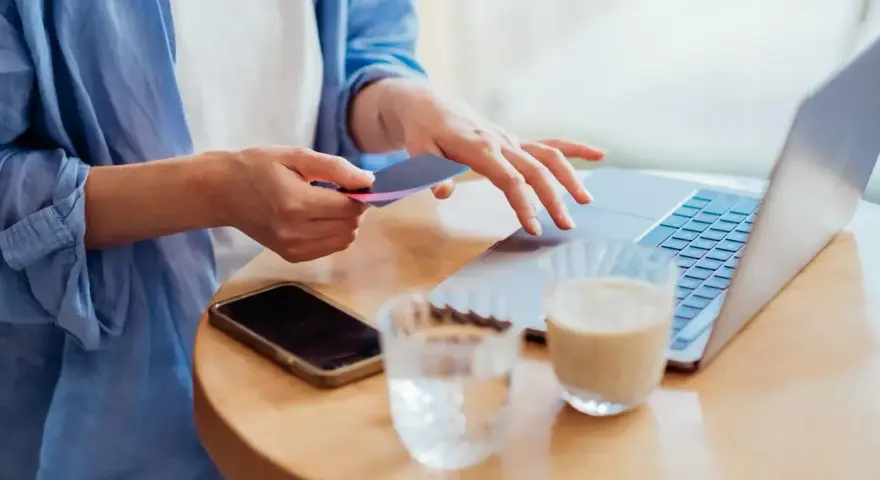 Kobieta dokonuje płatności kartą w internecie, wpisując dane na laptopie. Obok leży telefon i napoje.