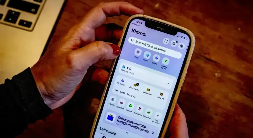 Dłoń trzyma smartfon z aplikacją Klarna. Czy Klarna jest bezpieczna? Aplikacja oferuje zakupy, płatności i zarządzanie zamówieniami.