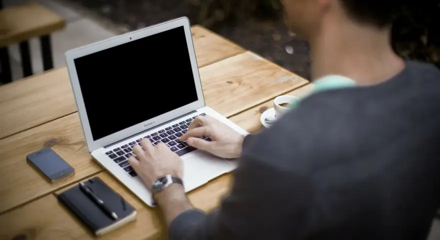 Mężczyzna pracuje na laptopie przy stoliku na zewnątrz. Obok leży telefon, notatnik i kawa. Może właśnie zarabia na **moneyo.pl**.