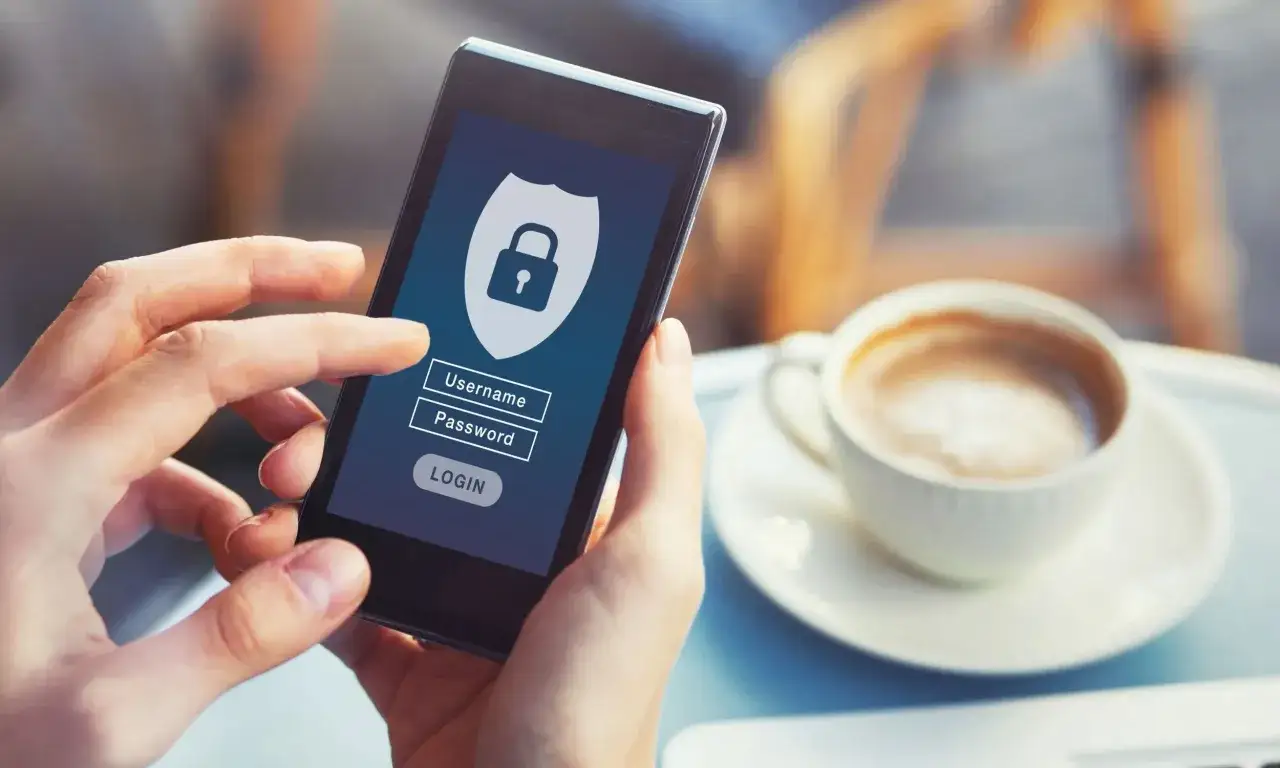 Dłoń trzyma smartfon z ekranem logowania do banku. Widoczny symbol tarczy z kł&oacute;dką, pola na login i hasło.
