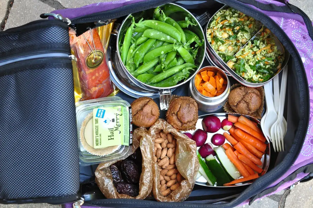 Kolorowa torba piknikowa pełna zdrowego jedzenia na drogę: fasolka szparagowa, sałatka ryżowa, marchewki, ogórki, rzodkiewki, daktyle, migdały, muffiny i tartę.